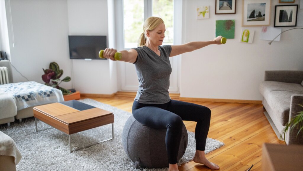 You Can Do This Entire Full-Body Dumbbell Workout Sitting Down woman sitting down on a gray stability ball doing a dumbbell workout in living room